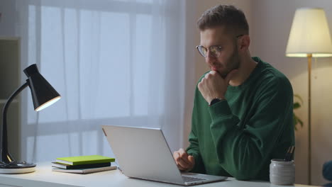 young-man-with-glasses-is-sending-message-in-online-chat-and-waiting-answer-using-internet-at-evening-at-home-male-portrait-with-notebook